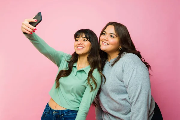 魅力的なヒスパニック系の女性の友人は スマートフォンでソーシャルメディアのために自撮りを笑っていますOピンクの背景 — ストック写真