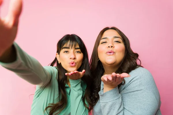 Personal perspective of a couple of best friends blowing kisses and taking a selfie together in front of a pink background