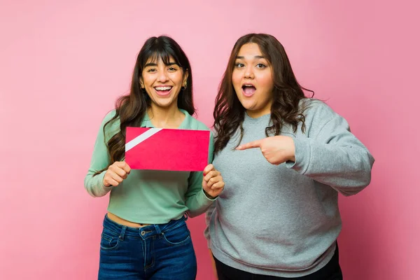 Happy hispanic women best friends pointing to a gift card and smiling against a pink studio background