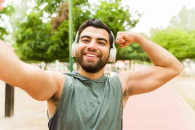 Strong happy man doing a bicep curl showing his arm muscles after exercising at the park looking cheerful
