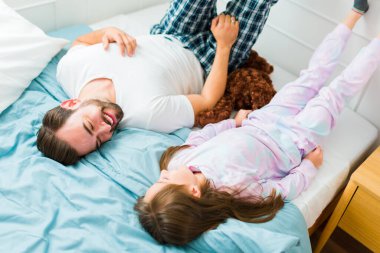 High angle of a loving relaxed father smiling and talking with her young daughter while in bed wearing pjs