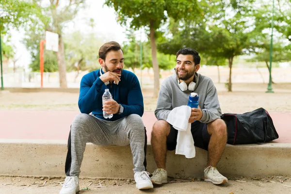Happy young men friends resting at the park while drinking water after finishing running and working out