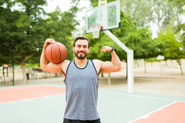 Strong happy caucasian man doing a bicep curl while smiling and ready to play basketball outdoors