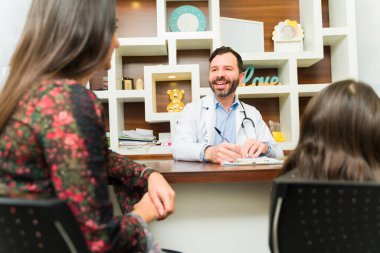 Neşeli çocuk doktoru mutlu görünüyor. Bir anne ve küçük çocuğuyla konuşurken gülümsüyor.