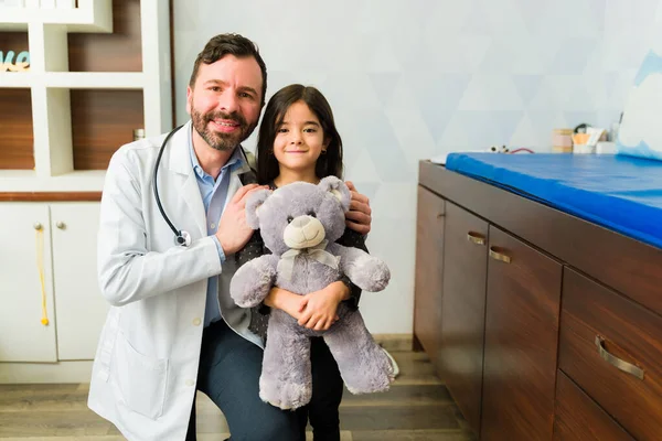 Cheerful caucasian doctor hugging a beautiful little child with a teddy bear coming to the doctor\'s office