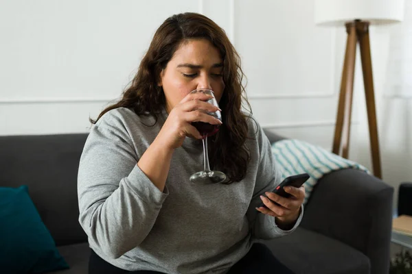 Overweight hispanic woman texting on the smartphone and drinking wine while looking sad and depressed