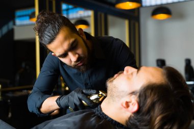 Mexican professional barber using a trimmer to shave the beard of a relaxed young man at the salon