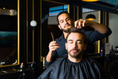 Hispanic man smiling while cutting the hair of a relaxed young man for a new haircut at the elegant barber shop