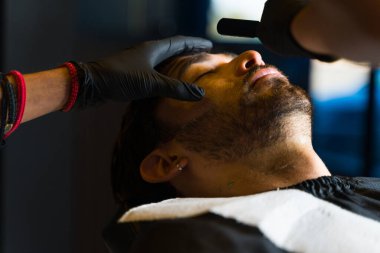 Attractive caucasian man relaxing while shaving his beard with a razor blade by a professional barber
