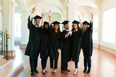 Attractive friends graduates looking excited showing their university diplomas wearing graduation gowns