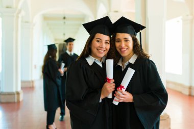 Beautiful happy women best friends posing with their college diplomas wearing graduation gowns