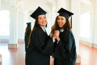 Women best friends with graduation gowns looking cheerful holding hands and celebrating getting university diplomas