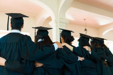 Young graduates seen from behind hugging in a line wearing graduation gowns and caps at university campus