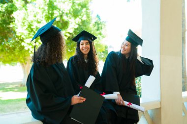 Cheerful best friends with graduation gowns holding their university diplomas and laughing on campus