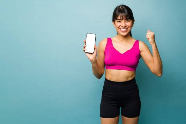 Excited sporty woman with an athletic body celebrating while showing her fitness app on the smartphone next to copy space