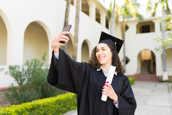 Attractive happy woman smiling taking a selfie with a smartphone wearing her graduation gown and holding her university diploma