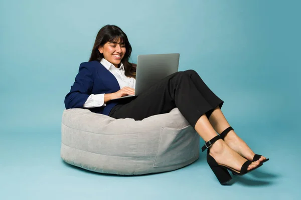Happy professional woman with formal clothes sitting on a puff chair and smiling while working and typing on the laptop