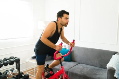 Determined young man with motivation using his stationary bicycle and exercising in his living room home
