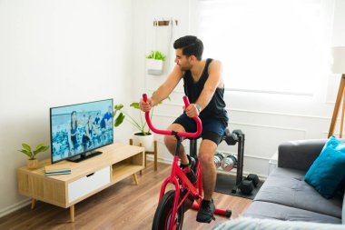 Smiling latin man riding his stationary bike at home while watching a workout video on the living room tv