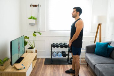 Side view of an active young man watching a home workout video and training with weights 