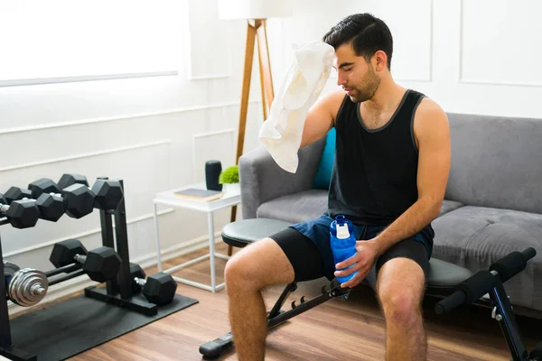Fitness young man sweating and using a gym towel holding a water bottle after exercising at home