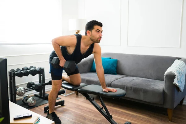 Hispanic young man in his 20s doing bench row exercises using weights while training in the living room