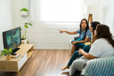 Latin female friends relaxing on the couch looking surprised while watching movies together in the living room