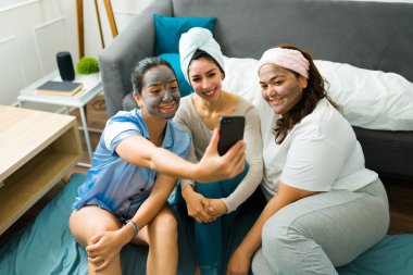 Smiling cheerful women and best friends taking a selfie while wearing face masks in their pajamas at home