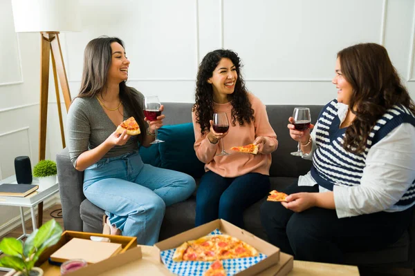 Smiling beautiful best friends laughing having a fun time while eating pizza and drinking wine sitting on the sofa
