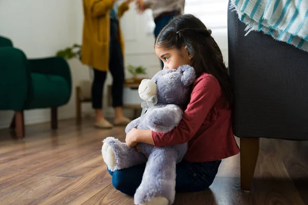 Sad young kid sitting alone hugging a teddy bear while looking scared about mom and dad screaming and fighting at home