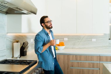 Çekici, rahat, gülümseyen, lüks beyaz mutfağının tadını çıkaran ve evde mutlu bir şekilde kahve içen bir adam.