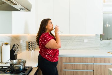 Gevşemiş şişman İspanyol kadın lüks bir mutfakta yemek pişirdikten sonra bir bardak şarap içmekten zevk alıyor.