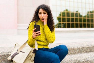 Kötü haber alırken şaşırmış gibi görünen genç bir kadın cep telefonundan sosyal medyaya bakıyor.