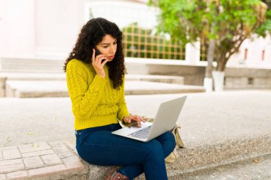 Genç bir kadın dışarıda çalışırken bilgisayarında yazarken akıllı telefondan iş görüşmesi yapmakla meşgul görünüyor.