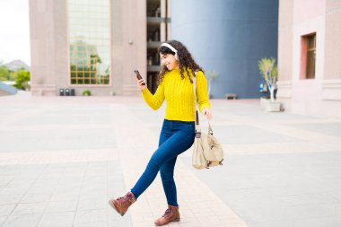 Genç ve mutlu bir kadının şehirde yürürken müzik dinlerken eğlenmesi ve akıllı telefonunu kullanması.