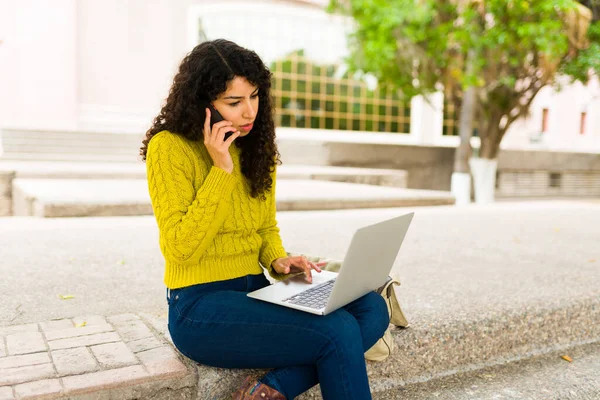 在户外远程工作时 年轻女人一边忙着用智能手机打电话 一边在笔记本电脑上打字 — 图库照片