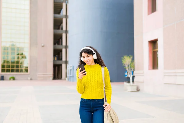 快乐的微笑的女人一边在城里散步一边用耳机听音乐 一边用智能手机发短信 — 图库照片
