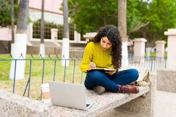 ノートに書いてノートパソコンを使いながら公園のベンチに座って遠隔作業をしている若い女性 — ストック写真