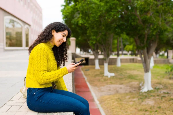 一个快乐而美丽的女人坐在公园外 一边微笑 一边用智能手机给朋友发短信 — 图库照片