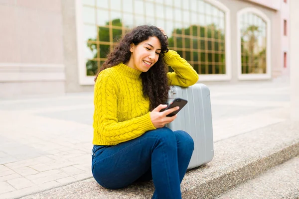 空港でスーツケースと一緒に旅行するのを待っている間 彼女のスマートフォンにテキストメッセージを送りながら 陽気な若い女性が笑っています — ストック写真
