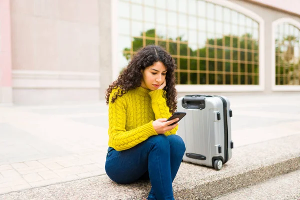 白种人的年轻女子带着手提箱坐在机场外等待出租车服务 同时使用一个搭便车共享应用程序 — 图库照片