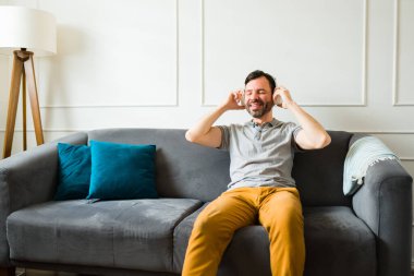 Mutlu beyaz adam neşeli ve rahat görünüyor. Kulaklıkla müzik dinliyor ve oturma odasındaki kanepede oturuyor.