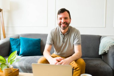 Çekici beyaz adam mutlu görünüyor. Oturma odasında laptopunu kullanırken gülümsüyor ve evden çalışıyor.