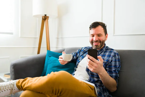 快乐的高加索人在客厅休息的时候 一边喝咖啡 一边用智能手机或社交媒体发短信 — 图库照片