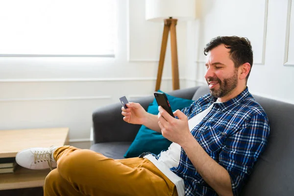 兴奋的男人一边在网上用智能手机购物 一边用信用卡付帐 在家里看起来很开心 — 图库照片