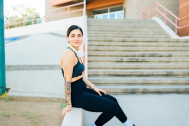 Neşeli genç bir kadın dışarıda antrenmanını bitirdikten sonra gülümserken kulaklık takıyor ve dinleniyor.