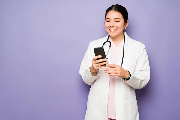 病院で医者として働きながら笑顔でスマホでテキストメッセージを送るアジア系の陽気な女性 — ストック写真