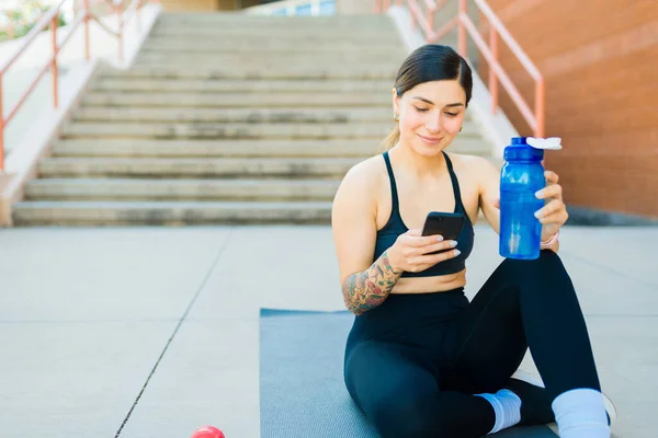 ハッピー魅力的な運動女性とともに水ボトルテキストメッセージで彼女のスマートフォン彼女のトレーニング演習を開始する前に — ストック写真