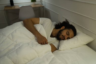 Kıvırcık saçlı latin adam rahat yatağındaki yorganın altında dinleniyor ve uyuyor. 