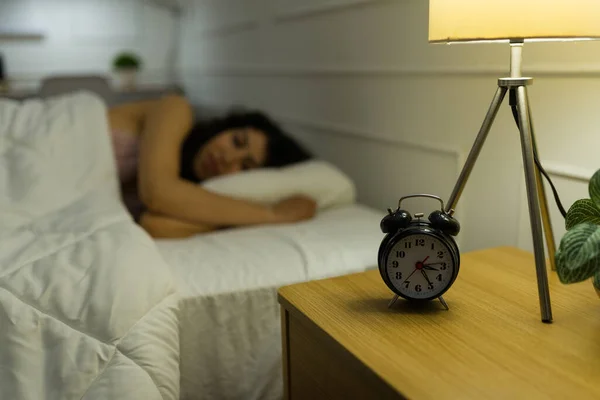 Focus on foreground of a night stand with an alarm clock next to restless young woman with insomnia trying to sleep in bed